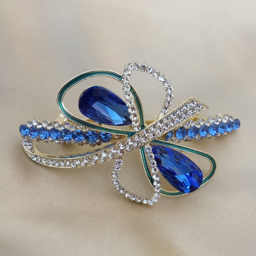Gold-tone hair barrette shaped like an abstract bow. Two large sapphire blue teardrop crystals sit at the center, surrounded by loops featuring teal enamel piping and sparkling clear pave rhinestones. Smaller blue crystals adorn the outer edges.