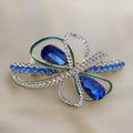 Gold-tone hair barrette shaped like an abstract bow. Two large sapphire blue teardrop crystals sit at the center, surrounded by loops featuring teal enamel piping and sparkling clear pave rhinestones. Smaller blue crystals adorn the outer edges.