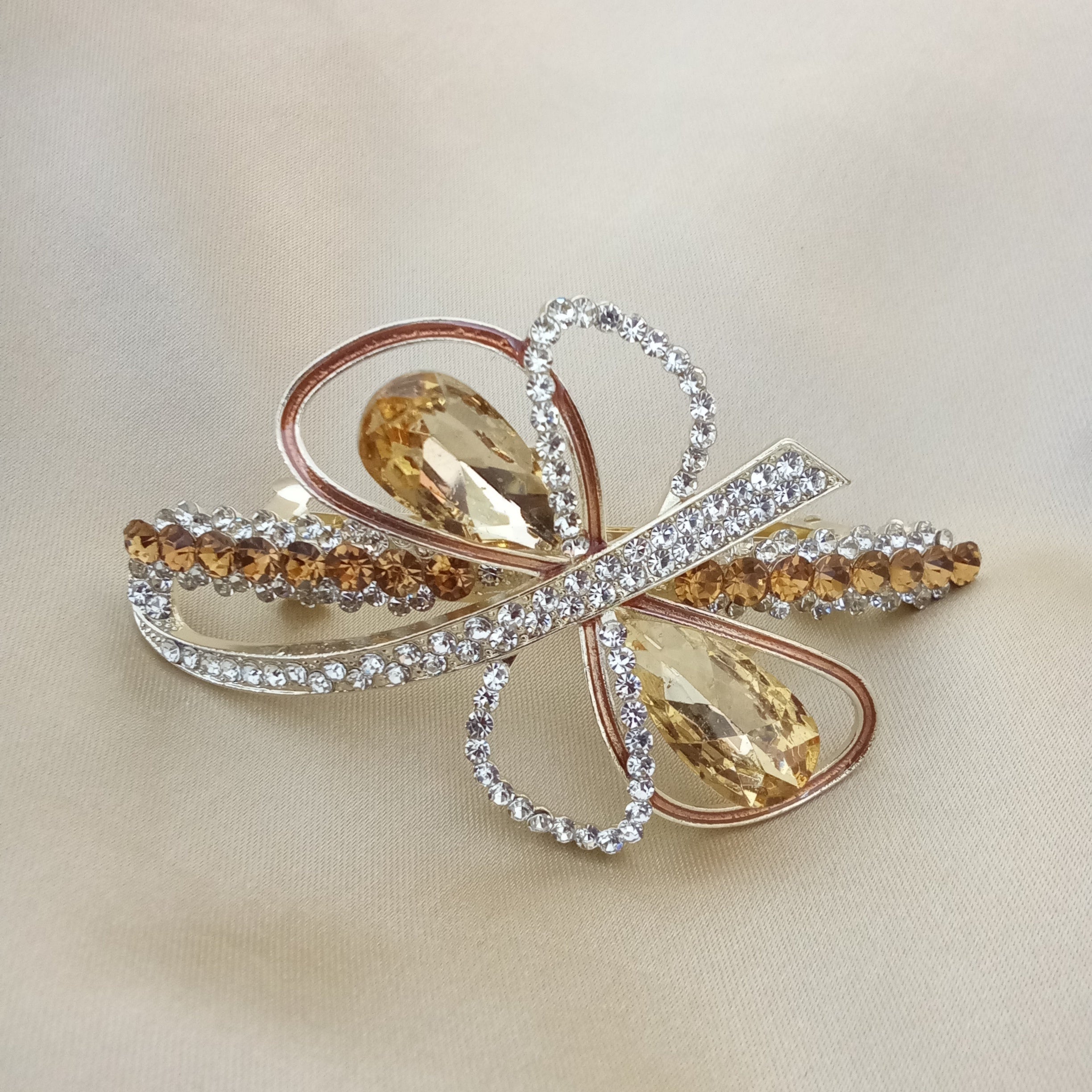 Gold-tone hair barrette shaped like an abstract bow. Two large brown teardrop crystals sit at the center, surrounded by loops featuring metallic copper piping and sparkling clear pave rhinestones. Smaller brown crystals adorn the outer edges.