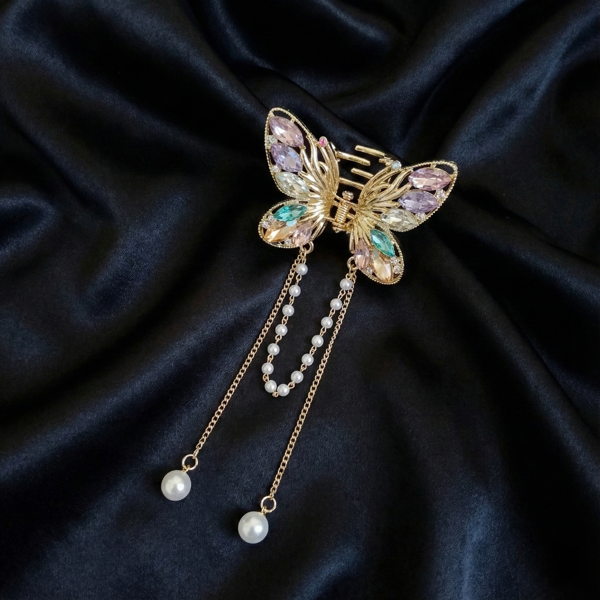 Gold-Tone butterfly hair claw clip with multi-colored crystals and dangling pearl tassel chains on a dark satin fabric background. Crated by Ladylike Accoutre