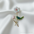Green and blue enamel hummingbird brooch perched on a gold-tone branch with an iridescent pearl flower and pink crystal center. Features intricate gold lines defining the bird's wings and tail.