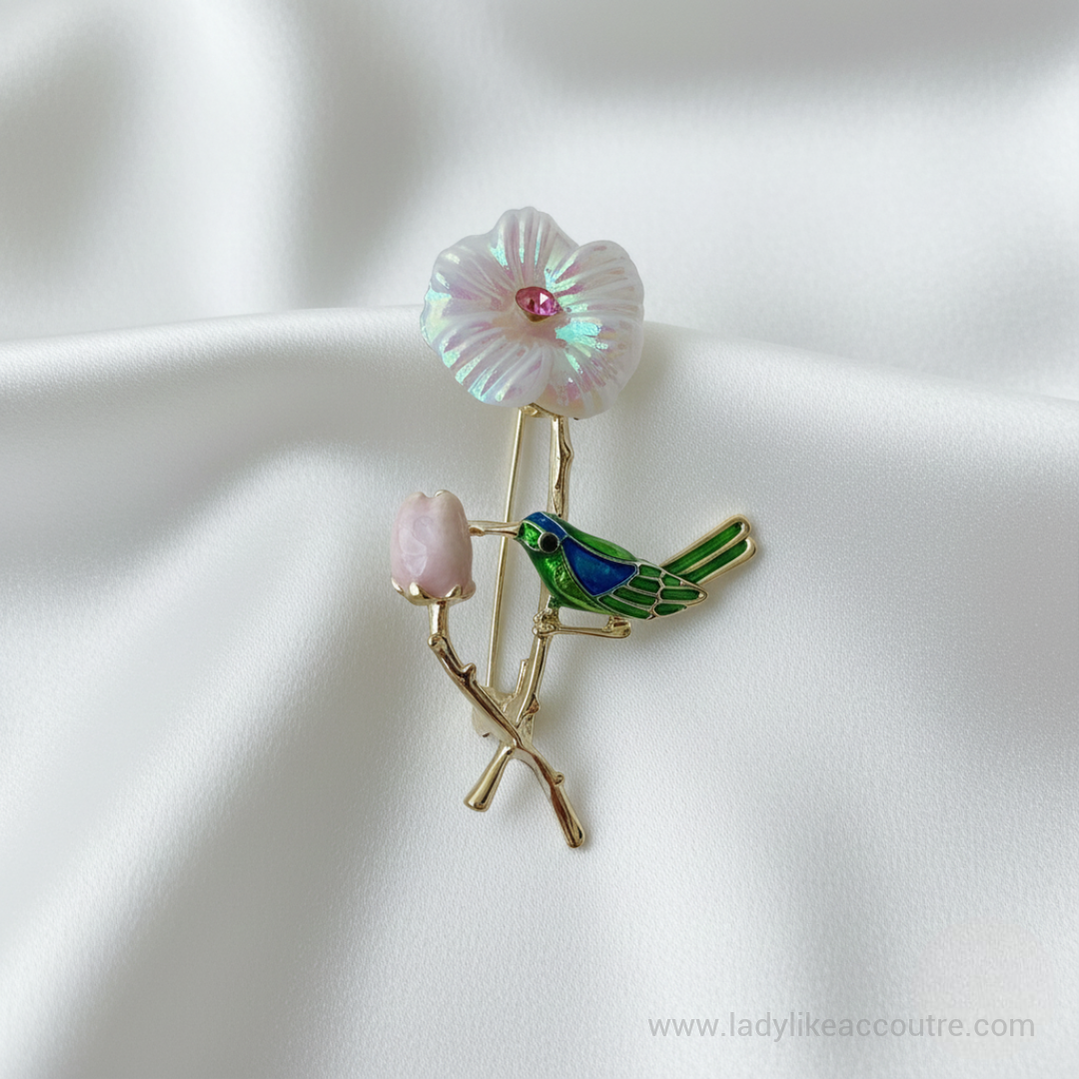Green and blue enamel hummingbird brooch perched on a gold-tone branch with an iridescent pearl flower and pink crystal center. Features intricate gold lines defining the bird's wings and tail.