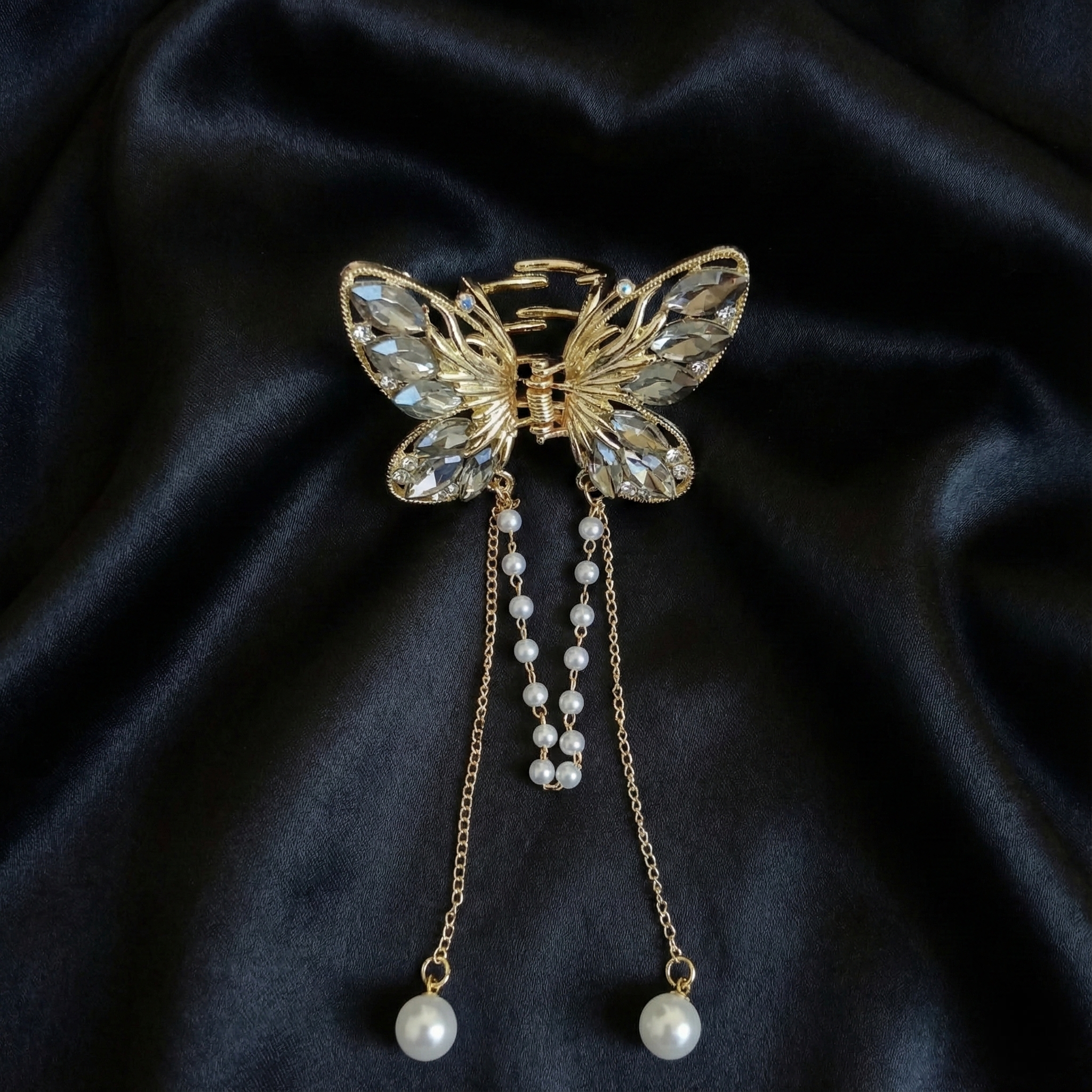 Gold-Tone butterfly hair claw clip with grey crystals and dangling pearl tassel chains on a dark satin fabric background. Crated by Ladylike Accoutre