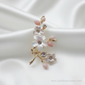Gold-Tone Pearl Flower Brooch Pin with crystal stone and pink bud, resting on a draped white satin background. The brooch features a winding gold-tone branch design adorned with three white flowers, each having pearlescent petals and a sparkling clear crystal center. Small, smooth pink buds accent the tips of the branches, and additional crystals are set along the stems, catching the light.