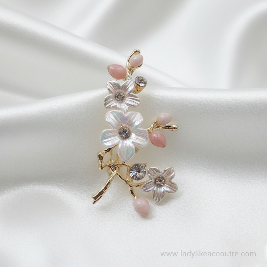 Gold-Tone Pearl Flower Brooch Pin with crystal stone and pink bud, resting on a draped white satin background. The brooch features a winding gold-tone branch design adorned with three white flowers, each having pearlescent petals and a sparkling clear crystal center. Small, smooth pink buds accent the tips of the branches, and additional crystals are set along the stems, catching the light.