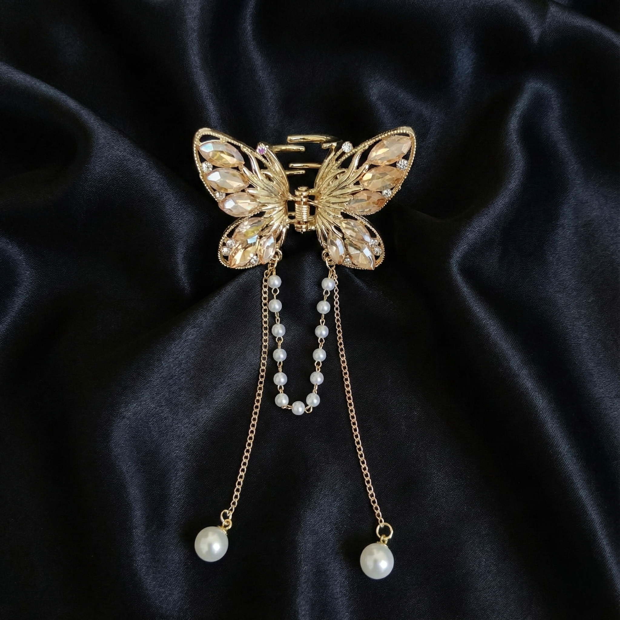 Gold-Tone butterfly hair claw clip with peach crystals and dangling pearl tassel chains on a dark satin fabric background. Crated by Ladylike Accoutre