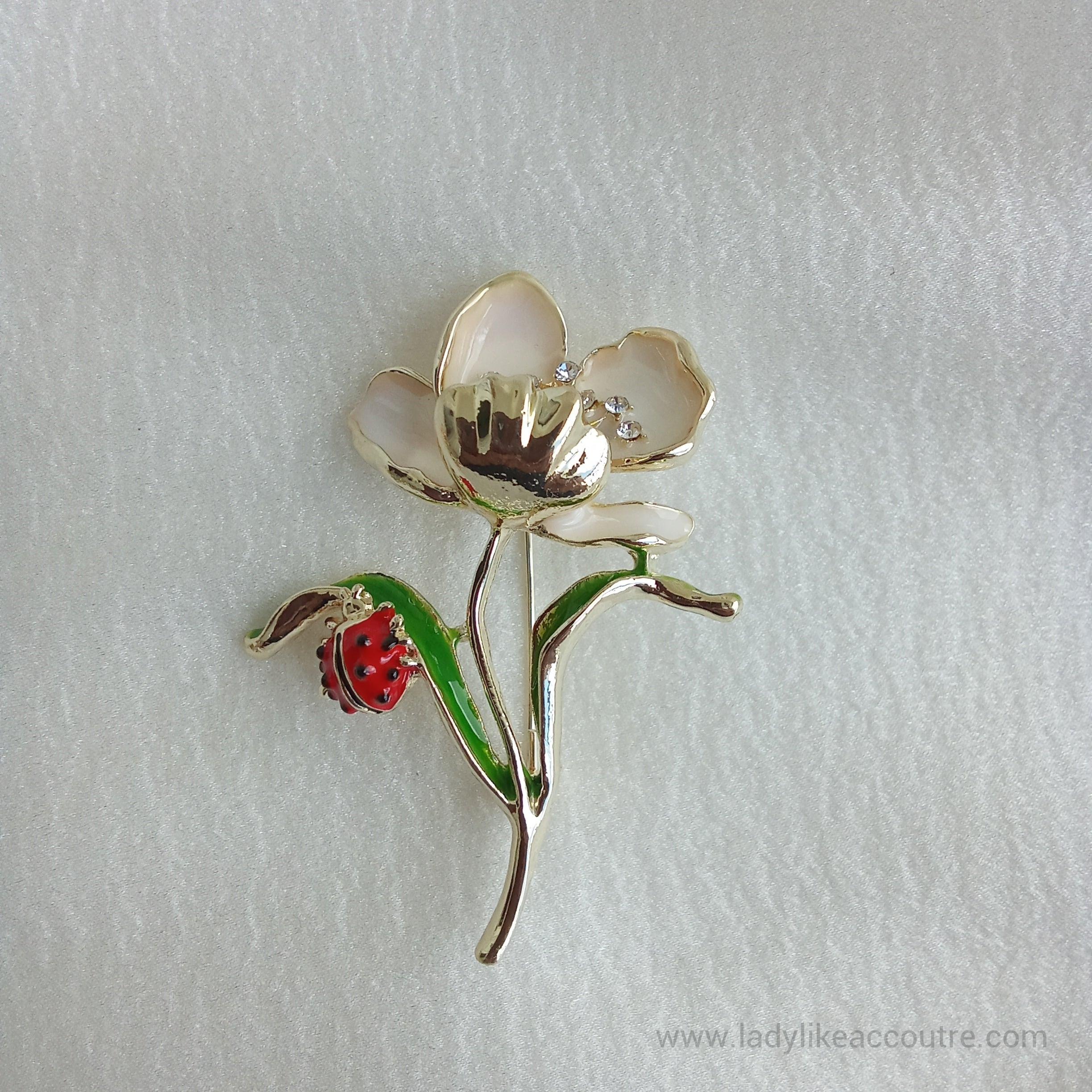 Gold-tone flower brooch featuring cream enamel petals and a sparkling crystal center. A vibrant green leaf branches off the stem, supporting a small, detailed red ladybug.