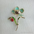 Gold-tone flower brooch featuring light turquoise blue enamel petals and a sparkling crystal center. A vibrant green leaf branches off the stem, supporting a small, detailed red ladybug.