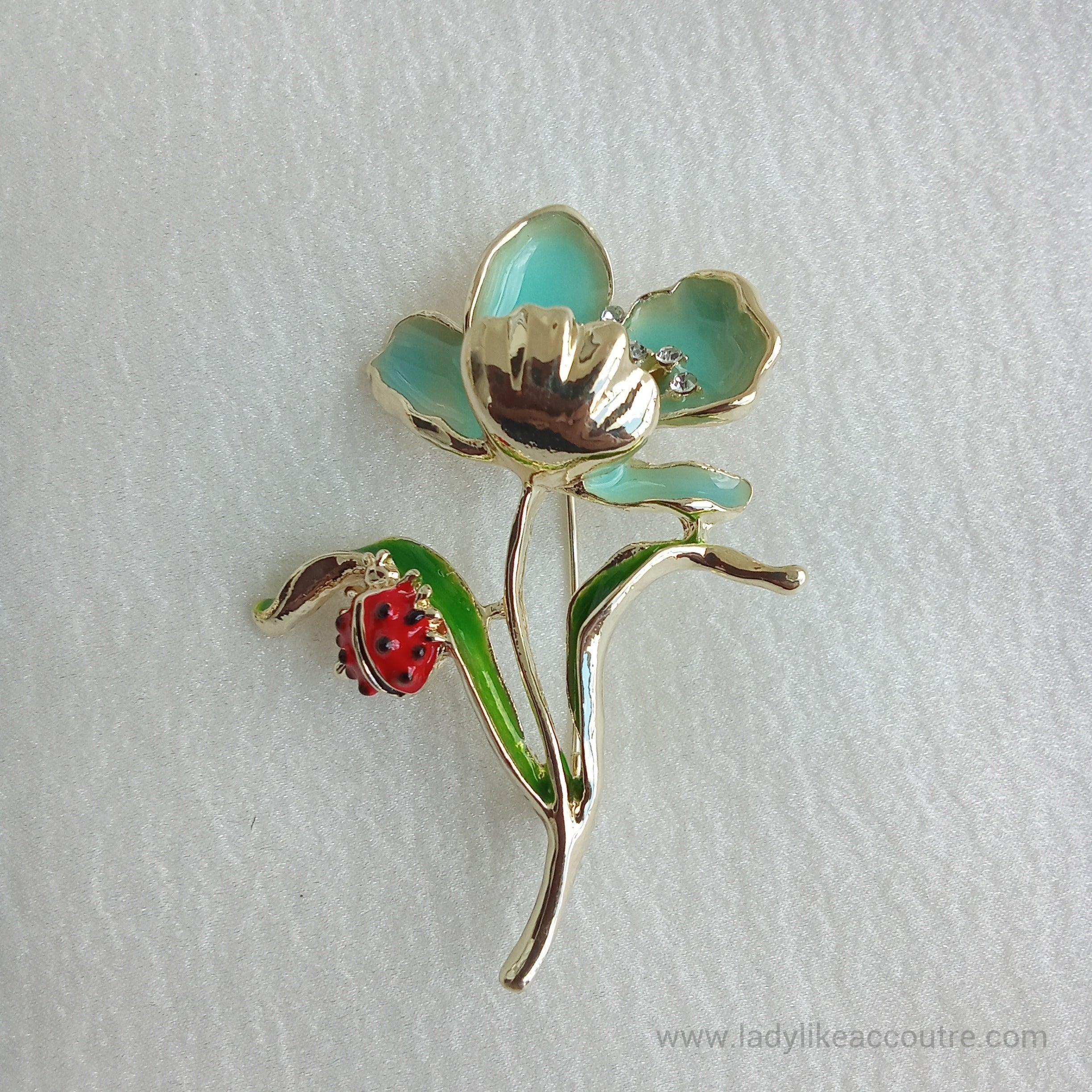Gold-tone flower brooch featuring light turquoise blue enamel petals and a sparkling crystal center. A vibrant green leaf branches off the stem, supporting a small, detailed red ladybug.