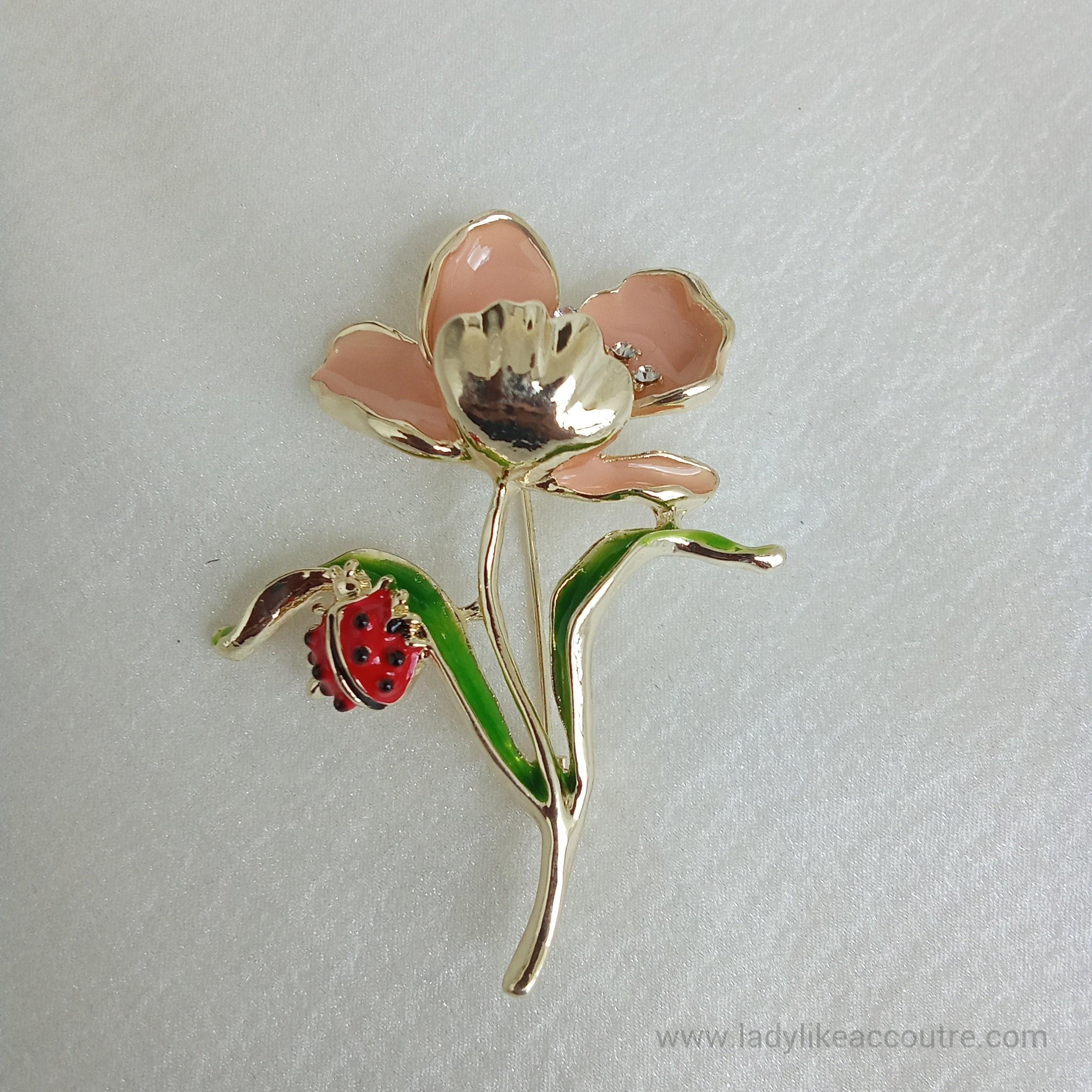 Gold-tone flower brooch featuring peach enamel petals and a sparkling crystal center. A vibrant green leaf branches off the stem, supporting a small, detailed red ladybug.