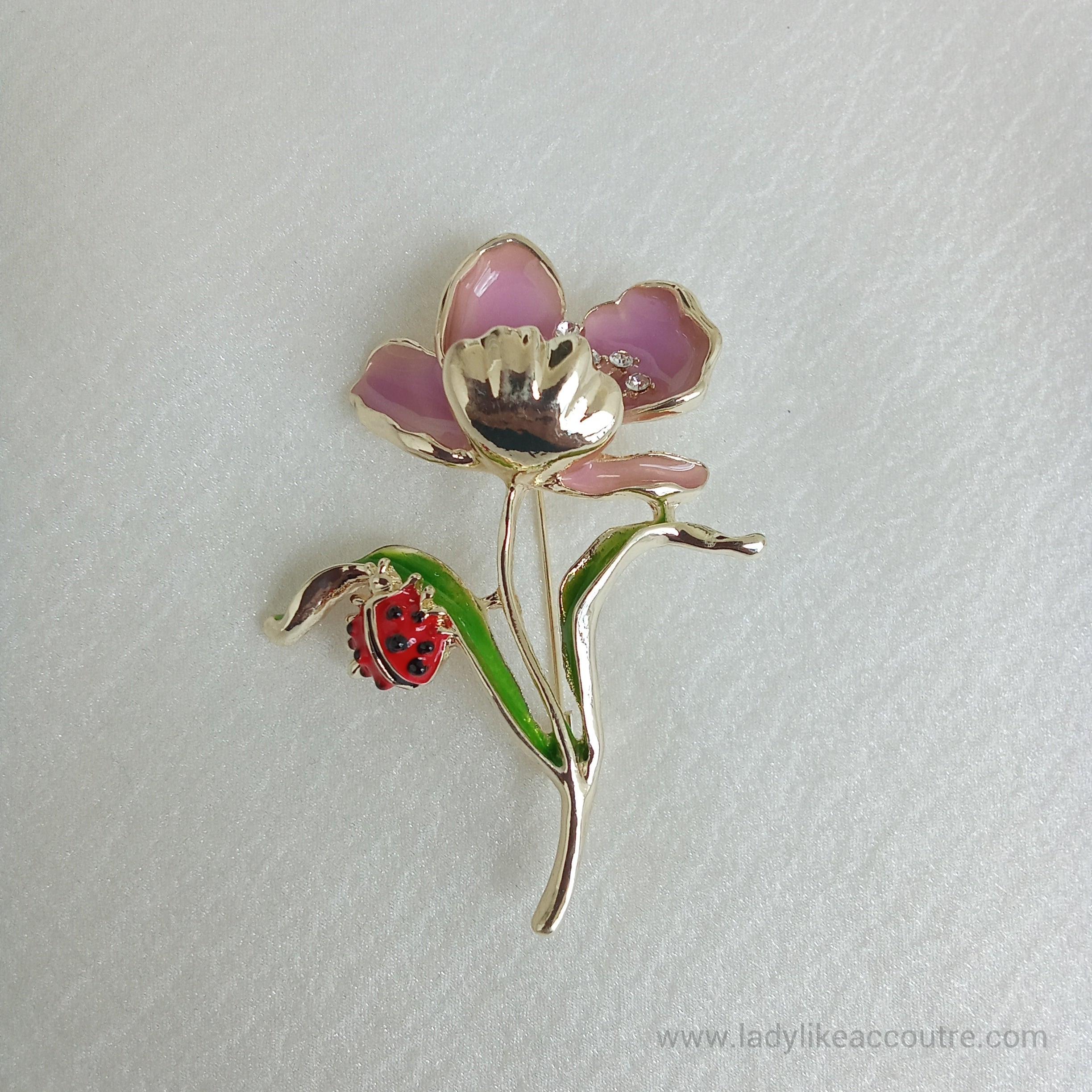 Gold-tone flower brooch featuring Lavender enamel petals and a sparkling crystal center. A vibrant green leaf branches off the stem, supporting a small, detailed red ladybug.
