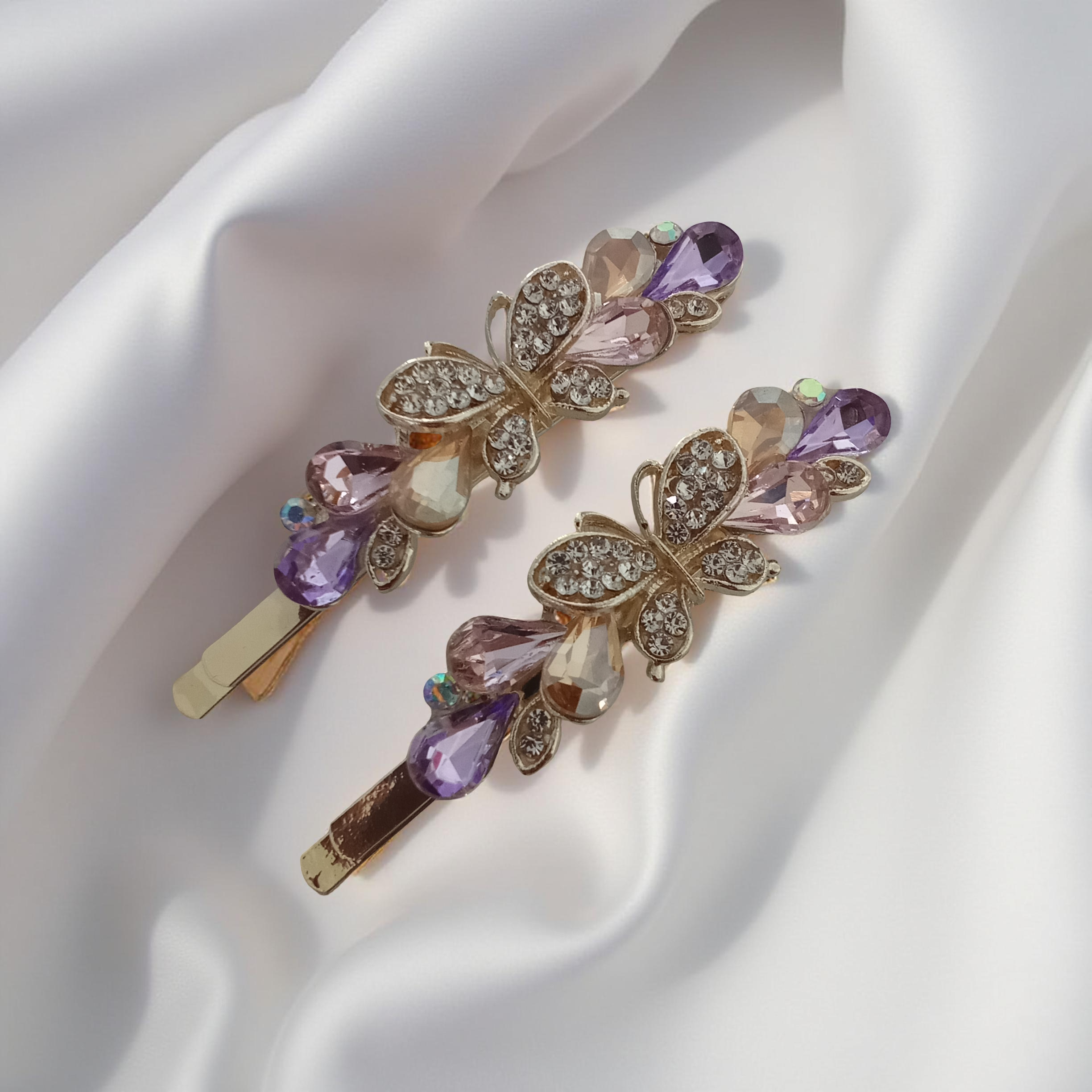 Pair of gold-tone butterfly hair pins featuring faceted multi-colored teardrop stones and pave crystals. Fancy hair slide for women displayed on a white silk background. Curated by Ladylike Accoutre.