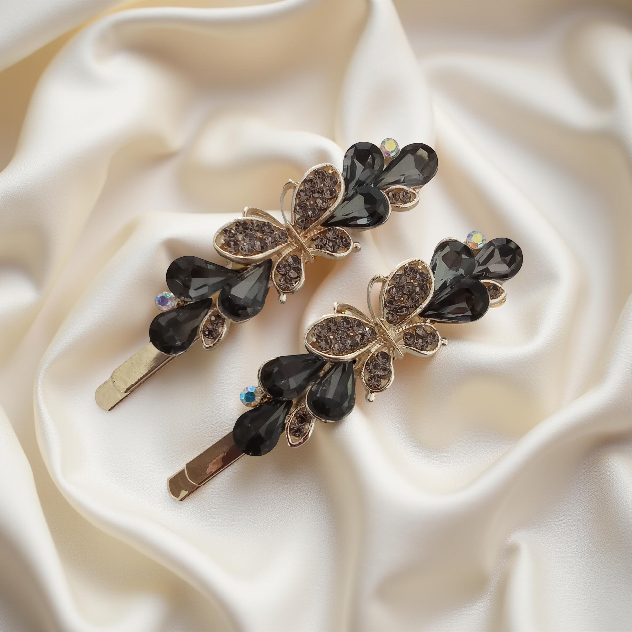 Pair of gold-tone butterfly hair pins featuring faceted black teardrop stones and pave crystals. Fancy hair slide for women displayed on a white silk background. Curated by Ladylike Accoutre.