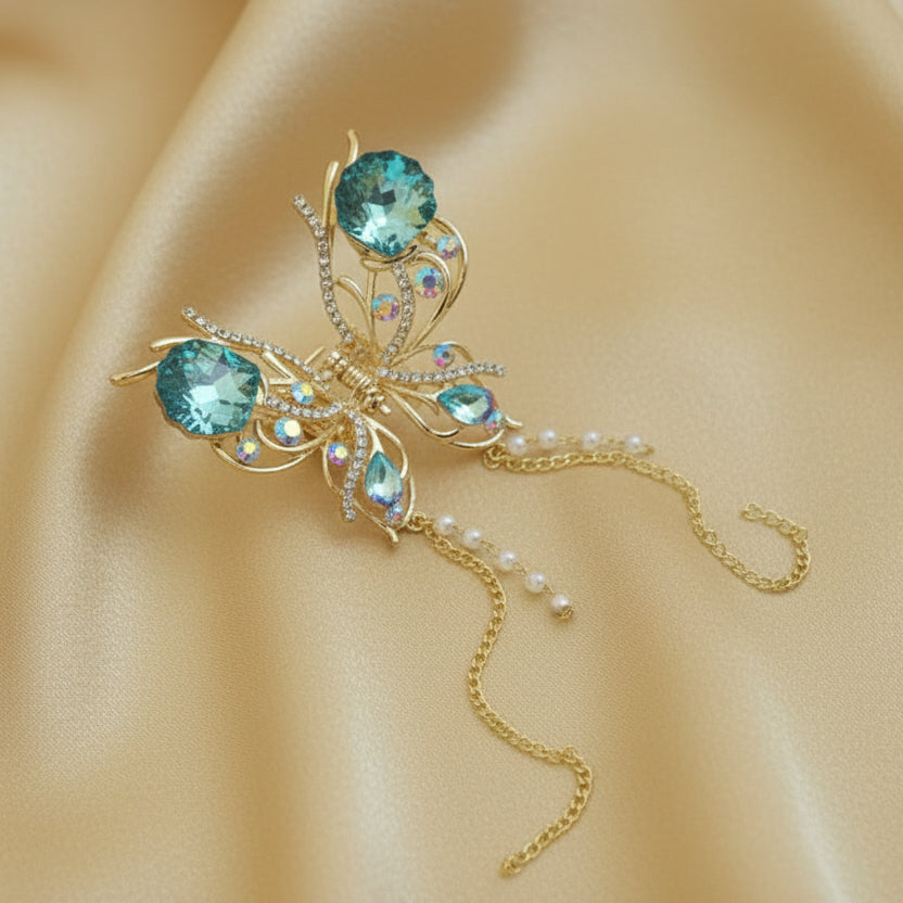 Large gold butterfly hair claw clip featuring aqua blue crystals and dangling gold chains with white faux pearls, set against a soft gold fabric background by Ladylike Accoutre