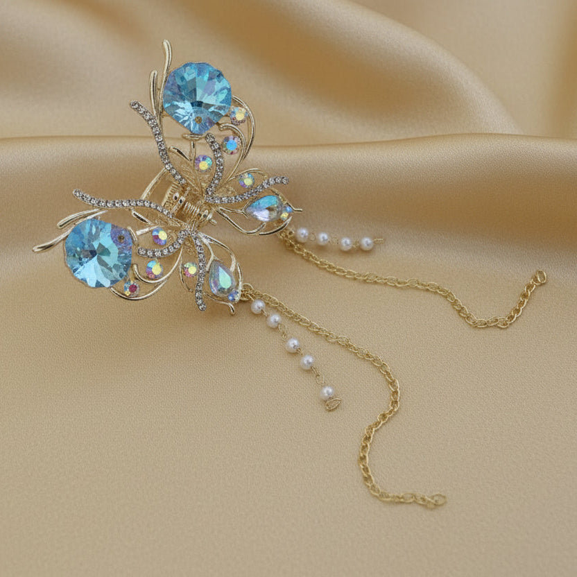 Large gold butterfly hair claw clip featuring white crystals and dangling gold chains with white faux pearls, set against a soft gold fabric background by Ladylike Accoutre