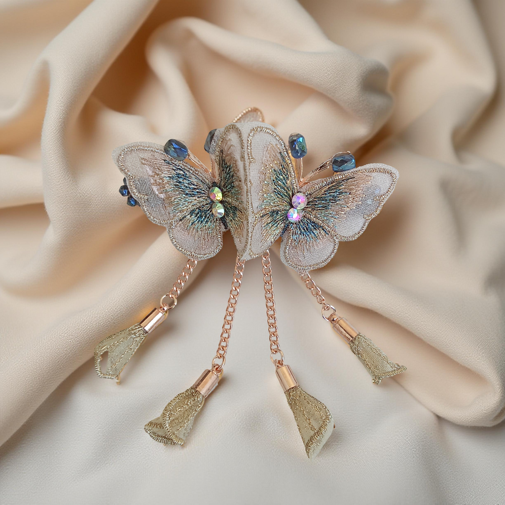 Twin embroidered butterflies in cream color on a rose gold-tone barrette hair clip with crystal accents and mesh charms, set against a soft cream background. Curated by Ladylike Accoutre.
