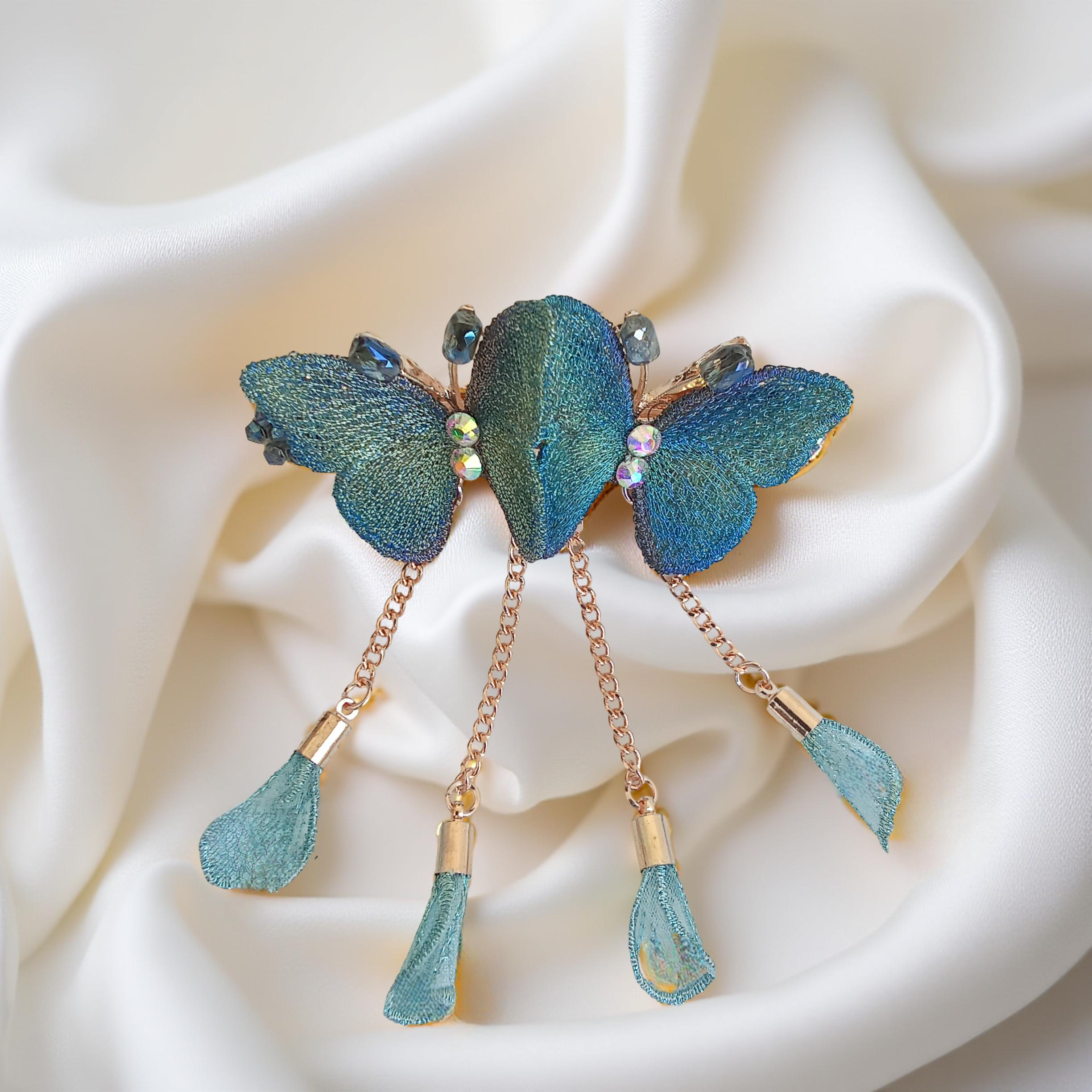 Twin embroidered butterflies in blue color on a rose gold-tone barrette hair clip with crystal accents and mesh charms, set against a soft white background. Curated by Ladylike Accoutre.