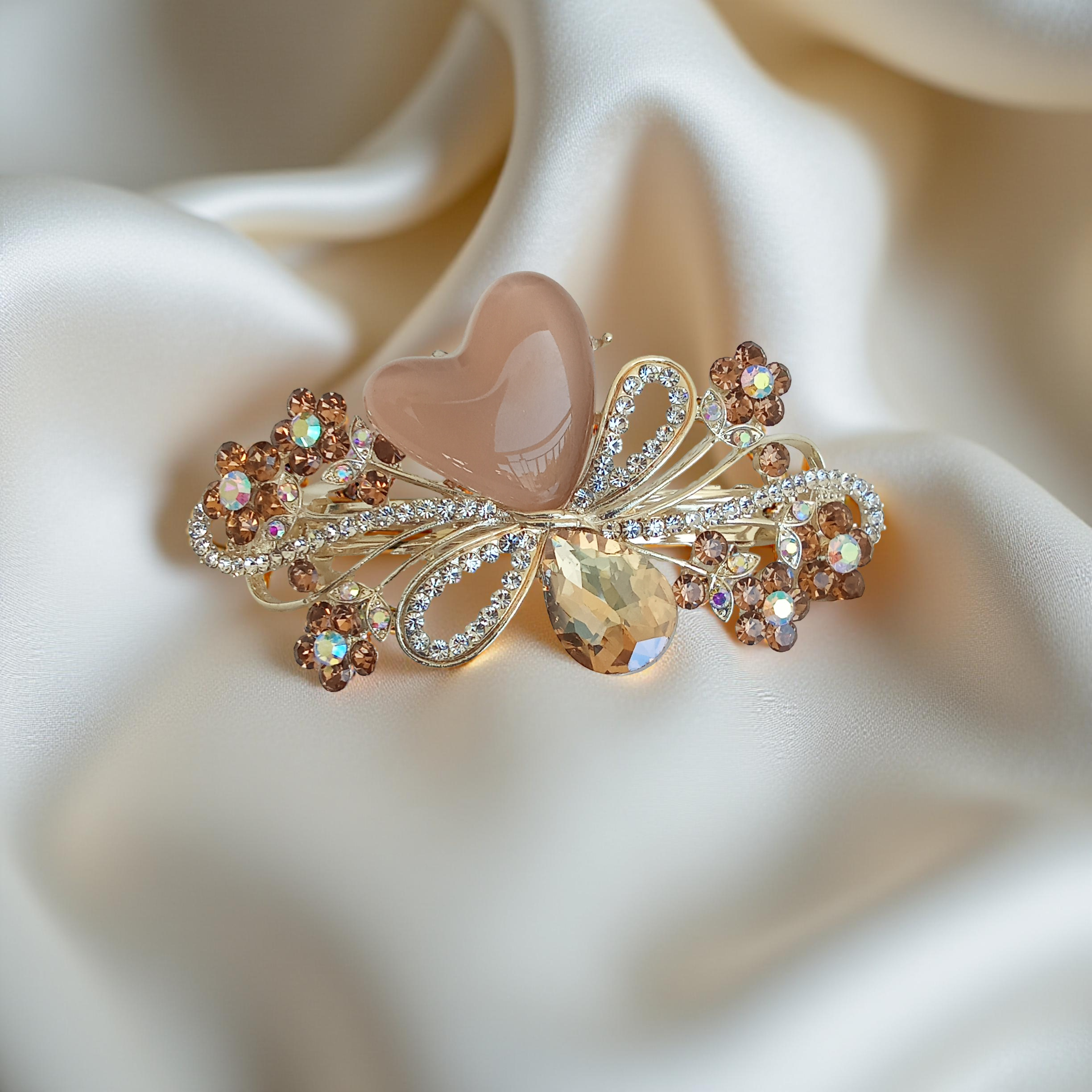 Ornate gold-tone floral barrette hair clip with a nude heart stone, amber crystal-encrusted flowers, and a smoky teardrop gem, displayed on a silky off-white fabric background. Curated by Ladylike Accoutre.
