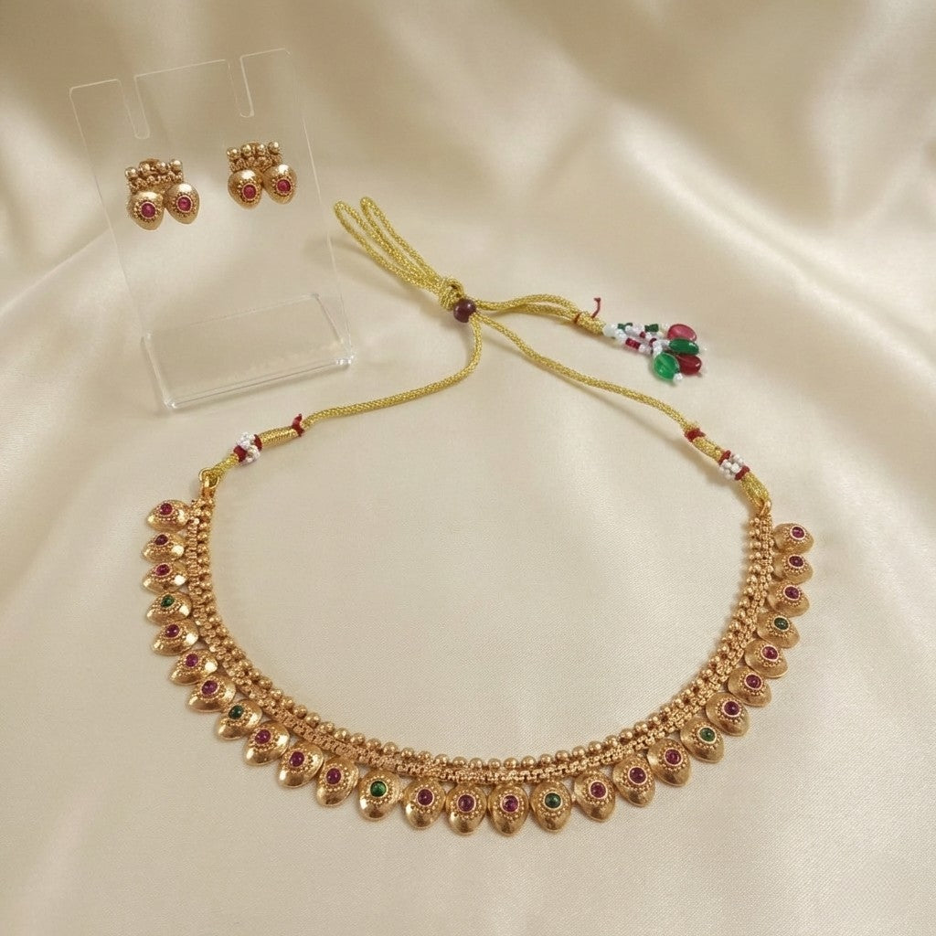 Gold-tone Mango Malai Kemp stone choker necklace set with ruby-red and emerald-green stones, adjustable dori, and matching stud earrings set on a cream shining background by Ladylike Accoutre.