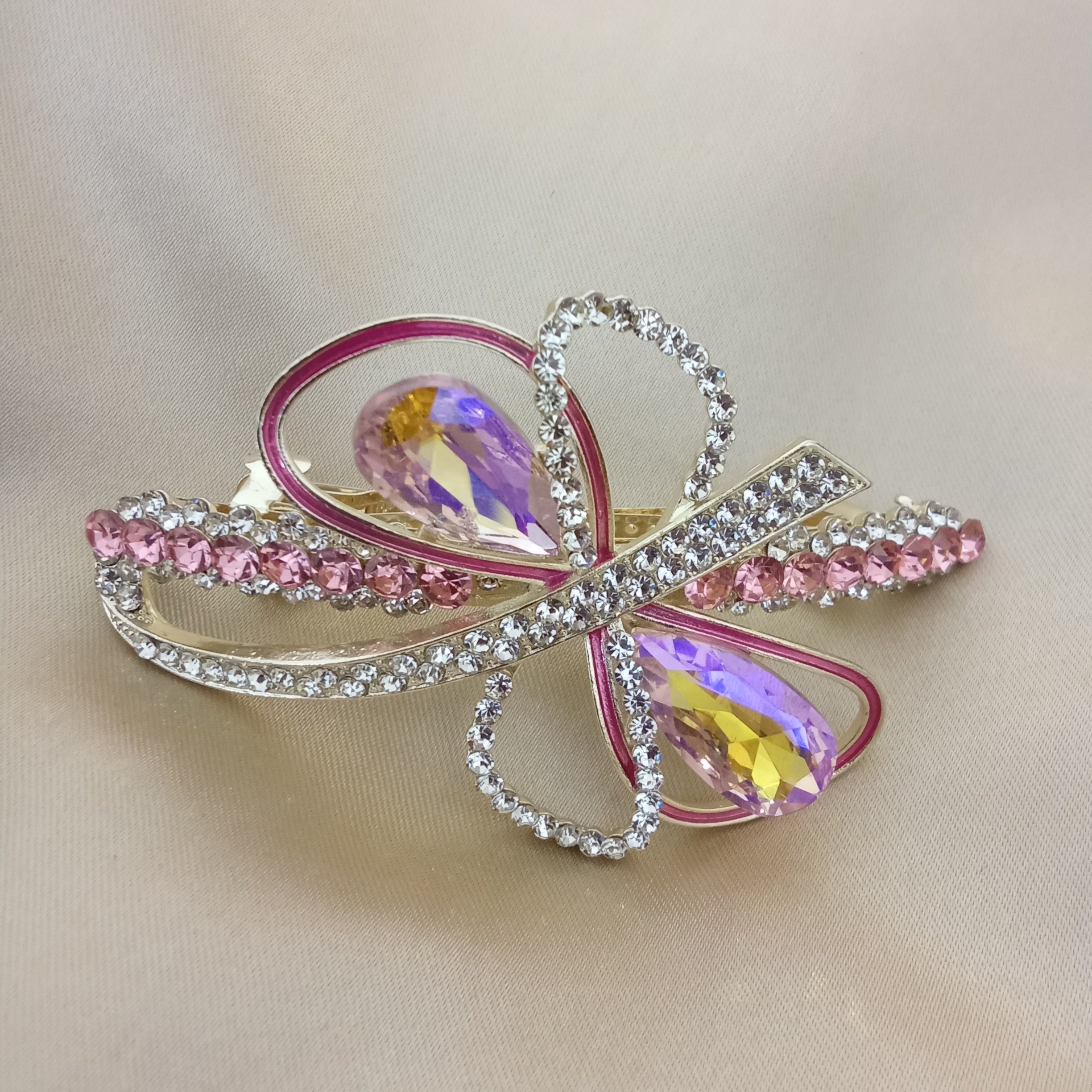Gold-tone hair barrette shaped like an abstract bow. Two large pink teardrop crystals sit at the center, surrounded by loops featuring pink enamel piping and sparkling clear pave rhinestones. Smaller pink crystals adorn the outer edges.