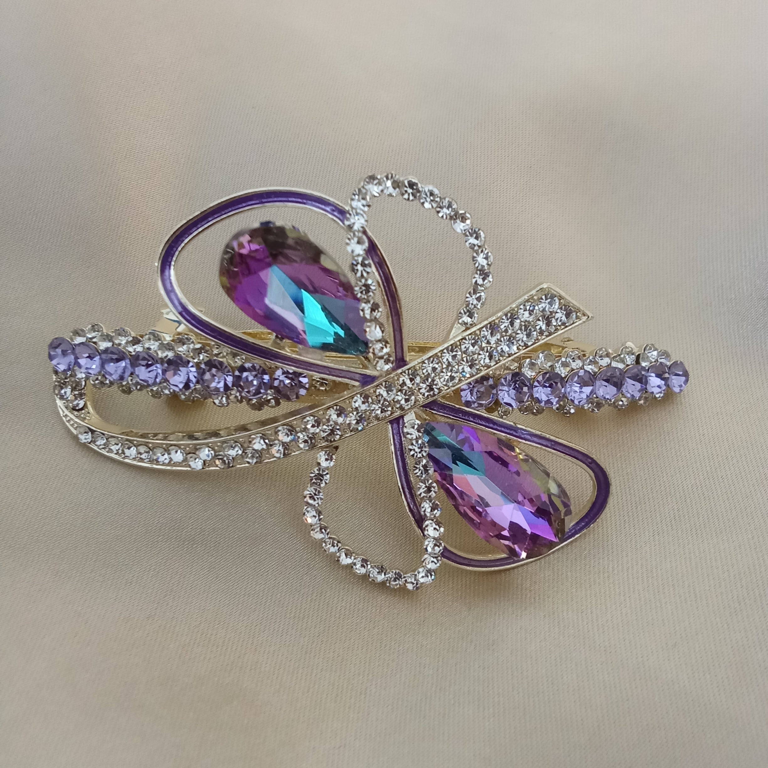 Gold-tone hair barrette shaped like an abstract bow. Two large purple teardrop crystals sit at the center, surrounded by loops featuring purple enamel piping and sparkling clear pave rhinestones. Smaller purple crystals adorn the outer edges.