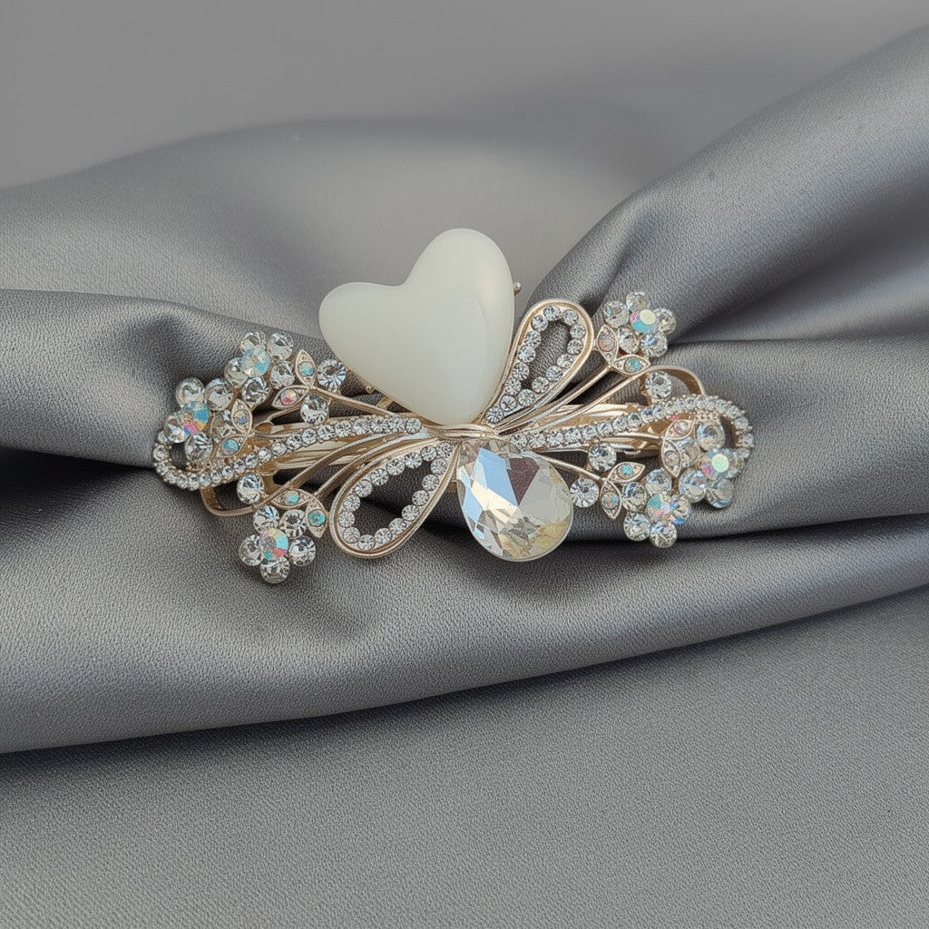 Ornate gold-tone floral barrette hair clip with white heart stone, crystal-encrusted flowers, and a smoky teardrop gem, displayed on a satin grey fabric background. Curated by Ladylike Accoutre.
